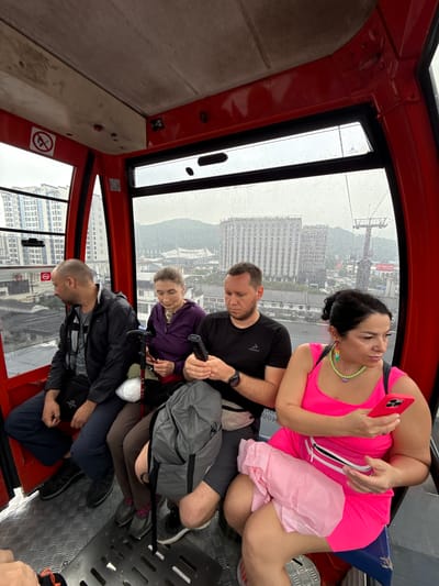 Inside a red cable car ascending above Zhangjiajie, China, four people are seated. A woman in a bright pink outfit and colorful necklace holds a phone, looking towards the window. A man in a black shirt is beside her, looking down. A younger person in a purple shirt sits next to him with walking sticks. A fourth person in a black jacket sits at the bottom. Outside, rain streaks the windows, obscuring views of the city's buildings and landscape.
