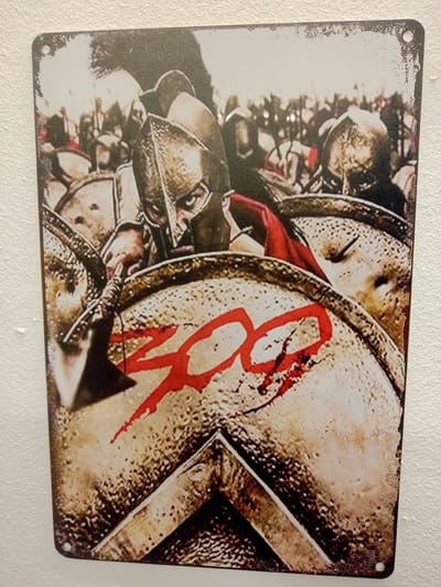 The image displays a metal sign featuring a graphic representation of warriors. In the foreground, a warrior wearing a helmet and armor is depicted, holding a spear and shield. Red lettering that reads "300" is superimposed on the shield. The background shows a multitude of other warriors in a formation. The sign is mounted on a textured white wall. No specific location in Nuremberg, Germany, is identifiable within the image.