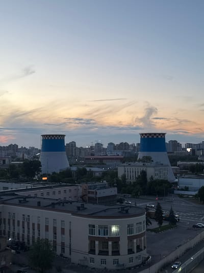 This is a high-angle, medium-shot, outdoor, daylight photograph of an industrial urban landscape in Bobrovo, Russia, taken during dusk. The sky is a pale blue with wispy clouds tinged with orange and yellow, indicating the setting sun. Dominating the mid-ground are two large, conical cooling towers, each with a distinctive blue band and a red and white checkered pattern at their tops. One of the cooling towers is emitting a plume of white steam. In the foreground and mid-ground, numerous buildings of various sizes and architectural styles are visible, suggesting a mixed-use urban environment with residential and industrial structures. Some of the buildings have dark, flat roofs. Trees are scattered throughout the scene, particularly in the lower left and mid-ground. A road network with several cars and a truck can be seen in the lower right portion of the image. The overall impression is a view of a city transitioning from day to night, with industrial elements playing a significant role.