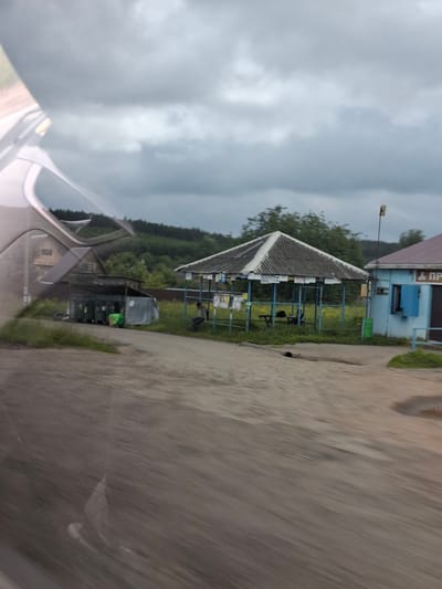 The image depicts a roadside scene in Забегалово, Russia, under a cloudy sky. In the foreground, a dirt road or pathway shows signs of motion blur. To the left, a weathered wooden building with multiple windows and a pitched roof is visible. Adjacent to it is an open-sided structure housing several large, dark green trash receptacles. Further to the right, a covered outdoor seating area with a corrugated roof and blue supports is set up, with tables and benches inside. Posters are affixed to the supports of this structure. Next to the seating area is a small, light blue building with a dark-colored entrance and a blue-shuttered window. A person is seated on a bench in front of this blue building. In the background, a dense line of trees forms a forest, with a hill beyond. The overall impression is of a rural or semi-rural location with functional, somewhat dilapidated structures.
