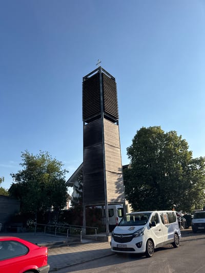 This is an eye-level, outdoor, full-shot photograph taken on a clear, sunny day in Höhenkirchen-Siegertsbrunn, Germany. The main subject is a modern church tower, constructed with a wooden facade and a metal frame, topped with a cross. To the right of the tower, a large, leafy green tree stands prominently. In the foreground, a white van with "Schulbus" (school bus) written on its side is parked on the asphalt road, adjacent to a sidewalk. A red car is partially visible in the lower left corner. The environment includes a paved sidewalk, a road, and greenery, suggesting a community area. The scene appears tranquil, with no people immediately visible. The lighting indicates it is likely late afternoon due to the long shadows cast.