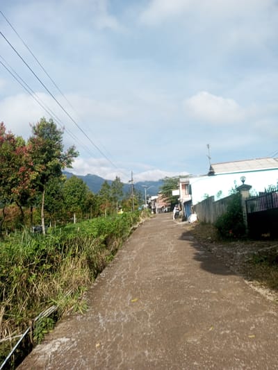 The image shows a paved road leading uphill through a rural or semi-rural area in Ein Chay Hnit Se, Myanmar. The road surface is worn. The left side of the road features a grassy embankment and a variety of trees, including trees with reddish leaves. Power lines are visible overhead. In the distance, mountains are visible. The right side of the road has a white building with a turquoise trim, a fence, and vegetation. A motorcycle is parked further up the road. The sky is partly cloudy.