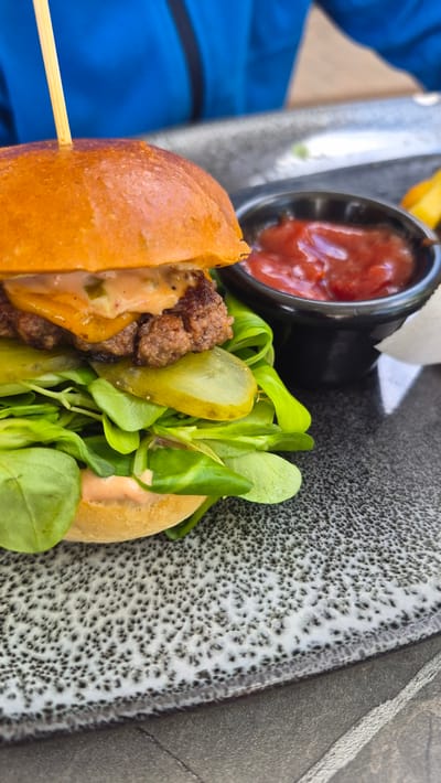 A close-up shot shows a hamburger with a wooden skewer in it. The hamburger has a golden-brown bun, a beef patty, cheese, lettuce, pickles, and sauce. In the background, there is a small bowl of ketchup and some fries. The food is served on a grey, textured plate. The setting appears to be outdoors, possibly a restaurant patio, given the bright lighting and the visible blue fabric of someone's jacket in the left of the frame. The overall impression is of a casual meal. There is no visible text in the image. The time of day appears to be daytime, and the weather seems pleasant.