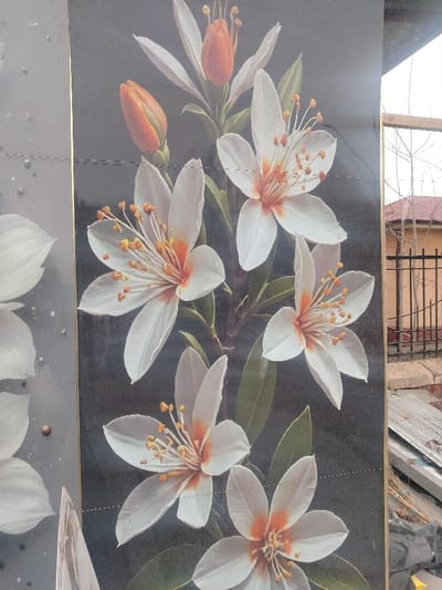 The image depicts a vertical arrangement of decorative panels, presumably installed outdoors in Balykchi, Uzbekistan. The dominant central panels showcase an artistic rendition of white flowers with prominent orange-yellow stamens and pistils, alongside two closed orange flower buds, all set against a dark grey or black background with green leaves. Faint horizontal seams delineate individual panels. To the left, a thin gold-colored vertical strip separates these from a partially visible panel featuring lighter floral elements on a grey background, accented with small gold-colored dots.
The right side of the image reveals an outdoor environment under an overcast, light grey sky. Bare deciduous trees with slender branches are visible in the mid-ground, beyond a black metal fence with vertical bars. A light orange-brown building with a dark roof is partially observable behind the fence. In the immediate foreground to the right, an accumulation of debris includes corrugated metal sheets, bundles of thin sticks or branches, and other unidentifiable refuse on a rough ground surface. The scene is consistent with a residential or semi-urban area in Balykchi, Uzbekistan. No individuals are present.
