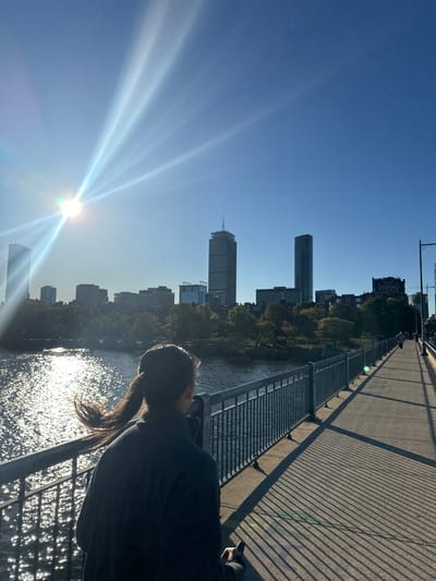 The image depicts a bright, sunny urban scene captured on the pedestrian walkway of the Harvard Bridge (Massachusetts Avenue Bridge) in Cambridge, Massachusetts, looking southeast towards the Boston skyline. In the foreground, a person with dark hair tied in a windswept ponytail, wearing a dark jacket, is viewed from behind, standing or walking and observing the distant cityscape. Their right hand holds a dark, rectangular object. To the person's left, the Charles River exhibits strong sunlight reflections, creating shimmering patterns. Metal railings separate the walkway from the water. Long, diagonal shadows from these railings extend across the concrete pathway on the right, indicating a low sun angle. The Boston skyline dominates the background under a clear blue sky, prominently featuring the Prudential Tower (tall, tapered, with an antenna) and 200 Clarendon Street (formerly John Hancock Tower), with other buildings and distant trees lining the riverbank. The sun, positioned in the upper left, generates significant lens flare and bright rays. Faint silhouettes of other pedestrians are visible further along the bridge on the right.