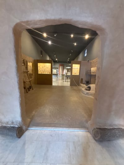 The scene is an interior view within a museum or exhibition hall in the city of Drama, Greece, observed through a light-colored, roughly textured archway. The archway's base incorporates rounded, dark-colored log-like elements. The immediate foreground floor is light-toned marble, featuring a thin blue illuminated strip at the threshold, which transitions to a smooth, light brown floor extending into the exhibition space. The ceiling is dark grey and equipped with numerous recessed spotlights, producing distinct lens flares.
On the left side, inside the exhibition area, a rustic display features multiple wooden pole structures leaning against a textured, light brown wall. Adjacent to this, an illuminated display case with an amber glow exhibits numerous small artifacts. On the right side, a low, curved, light-colored structure, resembling an ancient oven or hearth, is part of a display, positioned near another amber-lit display case containing additional artifacts. Further into the background, more exhibition space is visible, furnished with display tables or pedestals and additional cases. No individuals are present in the depicted scene.