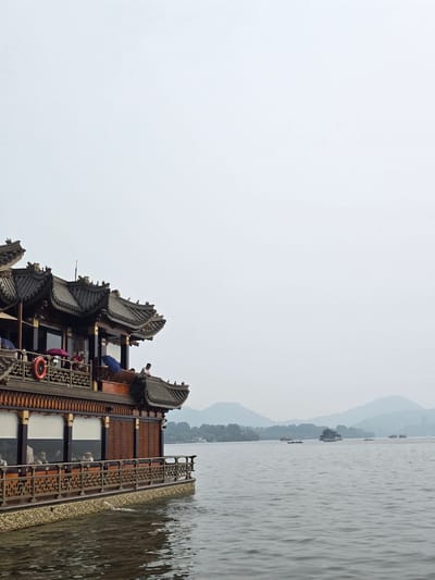 An ornate, multi-tiered traditional Chinese boat is docked on a calm body of water. The boat features dark wood construction with intricately carved railings and several levels of tiled roofs. Several people are visible on the boat's decks, some standing and looking out, while others are seated indoors behind large windows. One person stands on the upper deck, leaning over a railing. A life preserver ring is mounted on the railing of the second deck. The boat is likely a restaurant or tourist vessel. In the distance, across the water, a hazy landscape of rolling hills and other boats are visible. The sky is overcast and pale. The location is identified as a scene in Shangcheng District, China.