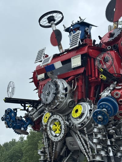 The image captures a close-up, low-angle view of a large, intricate sculpture made from recycled metal parts, likely located outdoors in a rural or park setting. The sculpture resembles a robotic figure or a complex mechanical creature, predominantly constructed from red and silver-colored industrial components.
Prominently featured are various gears, sprockets, bearings, and circular saw blades. One large, black and red saw blade is positioned towards the upper left of the frame, with a bicycle wheel rim mounted above it on a vertical pole. Multiple solar panels are attached to the red body of the sculpture, suggesting a theme of renewable energy or an artistic statement about technology.
Other visible elements include what appears to be a Gatling-gun-like apparatus with yellow accents, a wire fan, and numerous pipes, wires, and fasteners that contribute to the sculpture's detailed and complex appearance. The sculpture is a vibrant assemblage of repurposed materials, showcasing a creative and industrial aesthetic.
The background consists of a blurred expanse of green trees and a light gray, overcast sky. The overall impression is of a static, highly detailed art installation. The prompt specifies the location as Палыкавіцкі сельскі Савет, Belarus, indicating this is a public or private art display within that region. There are no people visible in the image.