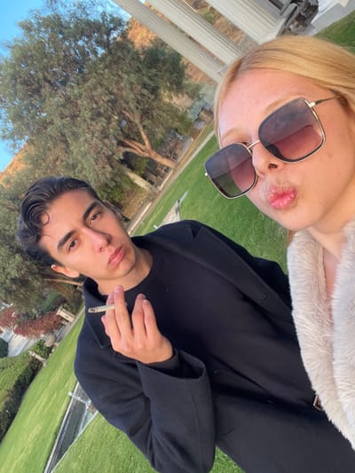 A selfie shows a blonde woman with sunglasses and a fur collar kissing the camera, and a man with dark hair holding a cigarette. The scene is outdoors, with a tree and building visible in the background, along with a well-manicured lawn. The man wears a black jacket and shirt. The woman's sunglasses reflect the sky.