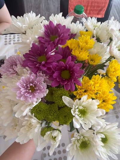 An overhead, close-up shot shows a colorful bouquet of chrysanthemums arranged in a white bucket. The flowers are in various stages of bloom and include white, yellow, purple, pink, and green varieties. A person's arm and hand are visible in the lower left corner of the frame, holding the bucket. In the background, an orange towel and a clear plastic bottle with a green cap are visible. The scene appears to be indoors, possibly in a home or office setting, with a table covered with a white cloth in the foreground. The image was captured in Omaha, United States of America.