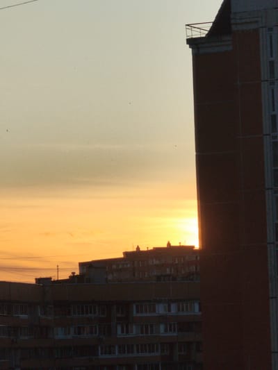 The image captures a dusky sky over the city of Korolyov, Russia. A tall, dark red brick building dominates the right side of the frame, partially obscuring the setting sun which casts a warm orange glow across the horizon. In the distance, silhouetted buildings with multiple windows suggest an urban residential area. The sky transitions from a pale yellow-orange near the sun to a muted grey-green at the top of the frame. Several small dark specks, possibly birds, are visible against the sky. The overall impression is one of a quiet evening in a city.