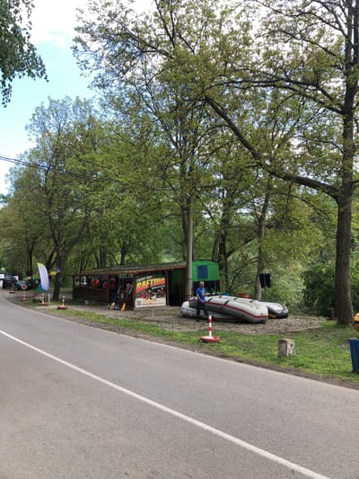 The image depicts a scene in Milanovo, Bulgaria, featuring a roadside establishment offering rafting services. Along a paved road, presumably leading into or out of the town, is a vendor’s booth or small shop displaying rafting-related equipment and promotional materials. A man wearing a blue shirt sits near an inflatable raft, partially obscured by a small green building. The rafting equipment for sale is at a booth underneath large trees. The booth is painted green and has a sign displaying information on rafting. The environment is characterized by greenery, including trees and grass bordering the road. A white stripe is marking the lane divider on the road.