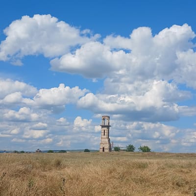 This is a photograph of a tall, dilapidated brick tower in a dry, grassy field under a bright blue sky with abundant white clouds. The tower, which appears to be an old church bell tower or watchtower, has arched openings at the top and shows signs of significant wear and tear, with exposed brickwork and crumbling sections. In the background, faint structures and trees can be seen on the horizon. The overall scene depicts a rural, somewhat abandoned landscape with a prominent, aging architectural element. The location is identified as Căzănești, Moldova.