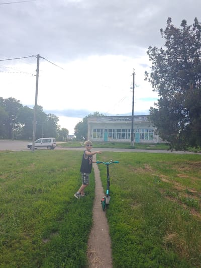 In an outdoor scene in Hadiach, Ukraine, a young boy with blonde hair and sunglasses stands on a dirt path next to a green and black kick scooter. He is wearing a black sleeveless shirt with a white logo and camouflage shorts. The boy is pointing to the right with his left hand, and his right hand is near the handlebars of the scooter. The scooter is positioned on the dirt path, facing away from the viewer.
To the left of the path, a patch of green grass slopes gently upwards. In the background, a white van is parked on a paved road. Further in the distance, a building with a light-colored facade and a green roof is visible. The building appears to be a store, with large windows and signage that reads "ГОСПОДАР" and "ТОВАРИ". Overhead, power lines are supported by wooden poles, and a large tree with dark green foliage stands to the right of the frame. The sky is overcast with gray clouds.