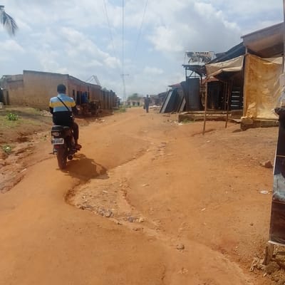 An unpaved, reddish-brown dirt road runs through a residential area in Ogbomosho, Nigeria. The road is uneven, with a ditch or erosion channel on the left side and scattered debris. On the right side of the road are several makeshift shops or market stalls with corrugated metal roofs and fabric awnings. One stall displays a large banner with a picture of a person. Buildings, some appearing to be made of mud bricks, line both sides of the road. Power lines stretch across the sky, supported by poles. In the foreground, a person rides a motorcycle away from the camera. The rider is wearing a blue and yellow striped shirt and dark pants. The motorcycle has a license plate visible. In the middle ground, another person walks away from the camera down the road. The sky is partly cloudy.