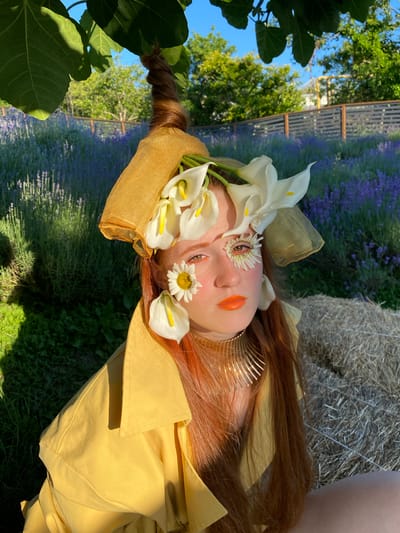 The image shows a person lying down, partially obscured by flowers and fabric. The subject's face is adorned with white calla lilies and a daisy, with vibrant orange lipstick. Their long, reddish-brown hair is styled into a braid on one side. They are wearing a yellow garment, possibly a coat or dress, with a gold comb in their hair. The background features a field of purple lavender, lush green trees, and a hint of a hay bale. The lighting suggests it is daytime, with clear skies and warm sunlight casting shadows. The overall aesthetic is ethereal and artistic, evoking a sense of nature and fantasy. The scene appears to be set outdoors, likely in a garden or rural area. The location context is Verkhneye Uchdere, Russia. No visible text is present in the image.