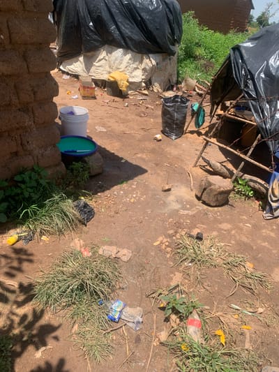 The image depicts an outdoor scene in Buga da Gindi, Nigeria. The ground is composed of dirt with some areas of mud and scattered debris, including plastic bottles, wrappers, and what appears to be dried vegetation. In the foreground, there is a patch of overgrown grass. To the left, a portion of a brick wall is visible. In the mid-ground, a white bucket, a green plastic basin, and a knife are present near the wall. A black plastic bag stands upright. Further back, a makeshift shelter constructed with branches and covered with black plastic sheeting is visible, with a wooden log and stones nearby. In the background, there are structures with corrugated metal roofs and dense green vegetation. The overall impression is one of a rustic and somewhat cluttered environment, suggestive of a living or working space.