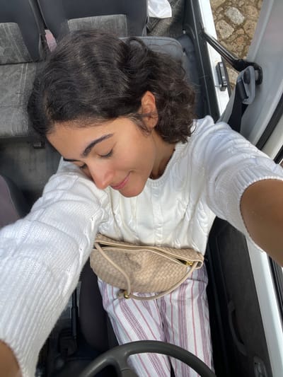 An overhead, slightly angled image captures a female individual seated in the driver's seat of a vehicle, parked outdoors in Vila Nova de Cerveira, Portugal. The subject has dark, wavy hair and is looking downwards with her eyes mostly closed, displaying a faint smile. She is wearing a white cable-knit sweater and light-colored pants with thin vertical red and white stripes. A light beige, woven fanny pack with a gold-colored zipper and a drawstring closure is positioned across her waist. A black steering wheel occupies the lower foreground of the image. Dark-colored seat upholstery is visible behind and to the left of the individual. The interior of the vehicle's white door frame, featuring a black hinge or latch mechanism, is visible on the right. Beyond the open vehicle door, a portion of cobblestone ground is discernible.