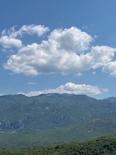 The image displays a panoramic view of a mountainous landscape under a bright blue sky dotted with fluffy white clouds. The foreground consists of dense green vegetation, likely trees and shrubs, with a slightly lighter shade of green in the immediate foreground. Behind this, rolling hills covered in a similar lush green foliage rise up. Further in the distance, a larger mountain range dominates the scene. These mountains are darker in color, a deep blue-green, and their rugged terrain is visible, with some lighter patches suggesting rocky outcrops or exposed areas. The peaks of these mountains are somewhat rounded and softened by distance. The sky above is a vibrant cerulean blue, with several large cumulus clouds scattered across it, casting soft shadows on the mountains below. The lighting suggests a sunny day, likely midday, with the sun high in the sky. There are no people or man-made structures prominently visible in the image, lending the scene a natural and somewhat remote feel. The overall impression is one of natural beauty and expansive, undeveloped wilderness.