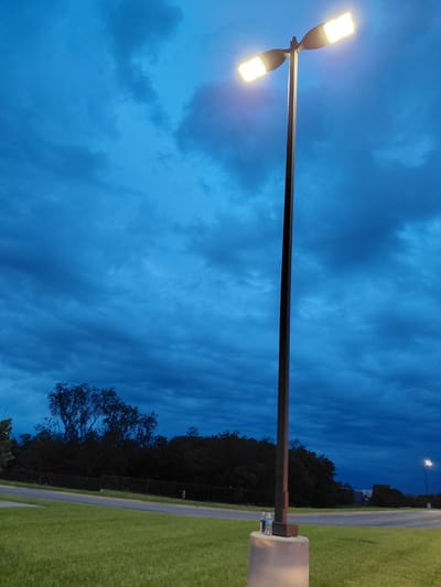 The image depicts an outdoor scene at dusk or dawn, with a deep blue sky filled with dramatic clouds. A tall, dark lamppost with two illuminated light fixtures stands prominently in the center-right of the frame. The lamppost is mounted on a concrete base. Two plastic water bottles are placed on top of the concrete base. In the foreground, a manicured green lawn stretches out. A paved road runs horizontally across the mid-ground, bordered by a dark line of trees and foliage. A black metal fence is visible behind the road. In the far distance, a distant light pole with a single light is visible. The overall setting appears to be a public space, possibly a park or a roadside area in Lexington, United States of America.