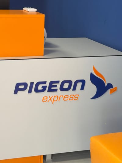 This indoor image, taken in Devin, Bulgaria, displays a commercial setting, likely a service counter or office. The central element is a large, light-colored vertical panel featuring the branding for "Pigeon Express." The logo, positioned at the top of the panel, is a stylized blue pigeon with an orange wing or tail detail, depicted holding a small orange envelope in its beak, symbolizing a delivery or postal service.
Below the logo, the company name is spelled out vertically: "PIGEON" in prominent, embossed blue capital letters, with "express" in smaller, orange, lowercase letters positioned alongside and slightly to the right of "PIGEON."
To the left of the branded panel, a section of a deep blue wall or ceiling is visible, creating a strong color contrast. In the lower left foreground, a bright orange cabinet or piece of furniture is present, with a small white electronic device or control panel mounted on its side. Another section of orange material, possibly a cushion or part of another fixture, is partially visible in the upper right of the frame. The scene is illuminated by artificial lighting, typical of an interior space, making the time of day and external weather conditions indistinguishable. No people or specific activities are captured in the image. The overall composition highlights the brand identity through its distinct color palette of blue, orange, and white.