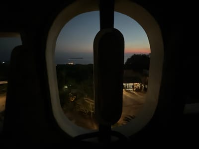 The image captures a view through a unique, oval-shaped opening, framed by dark, shadowy shapes that suggest an interior perspective. The scene outside depicts a twilight landscape with the sea in the distance, possibly at Karimunjawa, Indonesia. A faint glow on the horizon indicates the lingering light of sunset or sunrise, with a dark silhouette of a ship or large boat visible on the water.
Below the horizon, the land is covered in dark foliage, hinting at a tropical environment. Lights are visible on the land, suggesting human habitation or a settlement. In the mid-ground, a pathway or road leads towards buildings with illuminated interiors, where several figures can be seen through the windows, appearing to be customers or staff in a shop or restaurant. The overall atmosphere is serene and slightly mysterious, with the transition from day to night creating a muted color palette. The framing of the view through the oval opening adds a sense of curated observation, as if looking through a specially designed window or aperture. No visible text is present in the image.