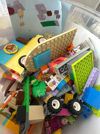 The image depicts a chaotic assortment of Lego bricks and building instructions spread across a table. The main focus is a collection of colorful Lego pieces, including various plates, bricks, wheels, and decorative elements. In the foreground, a yellow Lego vehicle is partially visible, adorned with wheels and a blue brick. Instruction manuals, featuring diagrams of Lego constructions, are scattered throughout the scene. The lighting is bright, casting shadows on the plastic surfaces of the Lego bricks. There are no people visible in the image. The background is blurred, but it appears to be an indoor setting with a white surface. The scene is set in Macclesfield, United Kingdom.