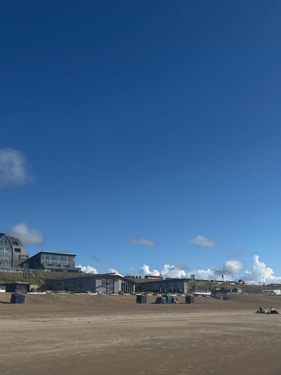 The image displays a sandy beachfront scene located in the coastal area of Overveen, Netherlands. The foreground consists of light brown, damp sand with visible tire tracks and ripples. In the midground, several dark, rectangular containers or bins are positioned on the sand in front of a series of modern, low-rise buildings, likely beach clubs or restaurants, featuring large windows and flat roofs, situated at the base of sand dunes. On the far left, a distinct multi-story hotel (NH Zandvoort), characterized by a curved glass facade and dark roof, is integrated into a dune. A dark flag on a flagpole is visible near the hotel. Towards the right side of the frame, three individuals are observed seated or lying on the sand. The background features rolling sand dunes with sparse vegetation and additional distant structures, some with red elements. The scene is illuminated by clear sunlight under a predominantly deep blue sky with scattered white cumulus clouds and a few small birds.