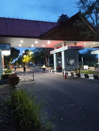 The media file depicts an exterior view of a gated entrance, likely a checkpoint or entrance to a facility, at dusk. The scene is set in Tambontunong, Indonesia. The sky is a deep blue, indicating twilight, and the lighting suggests artificial illumination from the overhead structure.
The main subjects include a gate, a small guardhouse or booth with a blue and white striped facade, and the paved road leading through the entrance. There are at least two individuals visible: one person in a white shirt and dark pants standing to the right of the road, appearing to be a guard, and another person on a motorcycle further down the road. Trees and foliage line the sides of the road and the background, suggesting a somewhat green or park-like setting.
Notable details include the red-tiled roof of the main overhead structure, the black and white checkered pattern along the road and barriers, and a red traffic cone placed near the gate. The overall atmosphere is calm and quiet, typical of the evening transition.
There is visible text on the guardhouse, appearing to be emblems or logos, one of which has the words "INSTITUT TEKNOLOGI" visible. Another sign with text is present on the right side of the road further in the distance. The weather appears clear, with no rain or strong wind suggested by the foliage. The time of day is clearly evening or dusk due to the sky color and lighting.