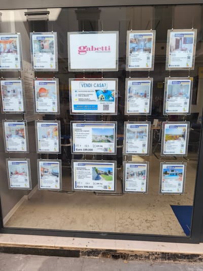 The image shows a shop window displaying real estate listings for gabetti Franchising in Livorno, Italy. The window is filled with numerous framed "FOR SALE" notices. Each notice contains a photograph of the property, along with details such as size, number of rooms, and price. The prices range from €55,000 to €690,000. A large "VENDI CASA?" (Sell House?) sign is prominently displayed, along with a QR code for more information. The shop window is set into a building facade, with the street visible in the reflection. The lighting suggests it is daytime. No people are directly visible in the scene.