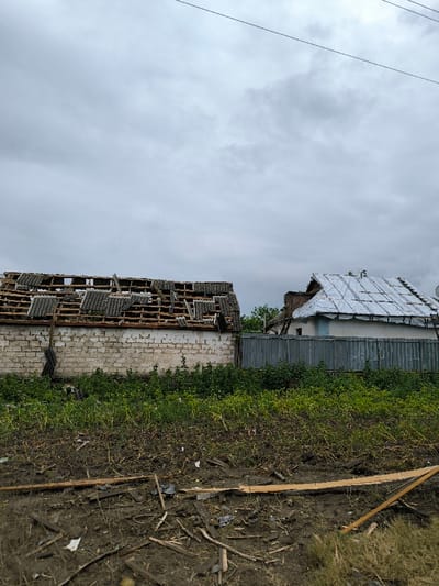 The image depicts a scene in Pysarivka, Ukraine, featuring two damaged residential or ancillary structures under an overcast sky. In the foreground, a disturbed patch of dirt is littered with debris, including numerous splintered wooden fragments and smaller pieces of dark material, possibly roofing. Two longer wooden planks lie diagonally in the center-right. Immediately behind this, a band of dense green vegetation separates the foreground from the buildings.

The structure on the left features a light-colored, possibly painted brick or cinder block wall. Its roof is severely compromised, with multiple dark gray corrugated sheets (likely asbestos or fiber cement) missing, broken, or dislodged, exposing the underlying wooden rafter framework. A dark, indeterminate object hangs vertically from the left side of this structure.

To the right, a corrugated metal fence, approximately 1.5 meters high, spans horizontally. Behind this fence, a second building is visible. Its wall appears light-colored, possibly white or light blue. The roof of this building is also significantly damaged, covered partially with a reflective, silvery material, possibly a tarp or corrugated metal sheets that are crumpled and dislodged. A small white satellite dish is affixed to its roof. In the upper portion of the frame, two thin, dark overhead wires stretch across the sky. No individuals, animals, or active movements are observed.