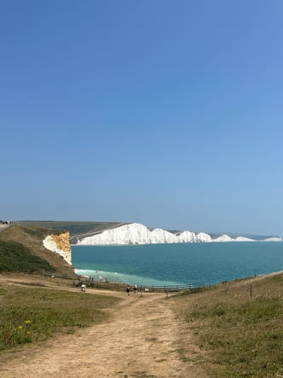 A wide shot captures the Seven Sisters cliffs near Seaford, United Kingdom, on a clear, sunny day. The bright blue sky dominates the upper half of the frame. The sea, a vibrant turquoise, stretches out in front of the majestic white chalk cliffs. The cliffs are irregular in shape, with some areas appearing to have eroded. In the foreground, a dirt path winds through dry, golden grass. A wooden fence runs along the edge of the path, separating it from a sloping grassy area. Several people are visible on the path and on the cliff edge, some walking and others standing. Two bicycles are parked near the fence. In the distance, the white cliffs extend along the coastline, gradually fading into a hazy horizon. The overall impression is one of a scenic coastal landscape with a few visitors enjoying the view.