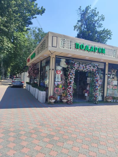 In Tursunzoda, Tajikistan, a flower and gift shop is captured on a sunny day. The shop, with its modern facade and large glass windows, is adorned with an elaborate archway of colorful flowers. Inside, large teddy bears and floral arrangements are visible. Outside the shop, a grey sedan is parked on the left side of the frame, facing away from the viewer. The pathway in front of the shop is paved with interlocking grey and pink tiles. The scene is framed by lush green trees and a clear blue sky, indicating an outdoor urban environment.