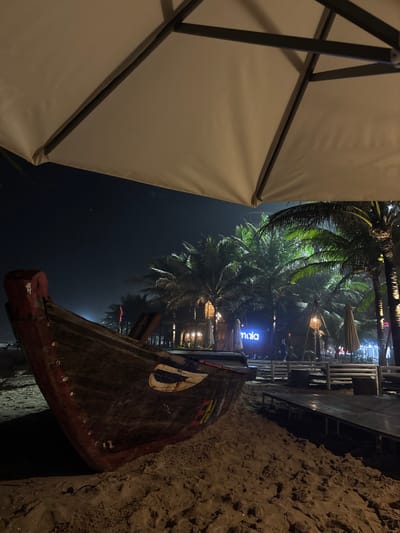 This nighttime image depicts a beach scene in Ngũ Hành Sơn Ward, Vietnam. The upper portion of a large, beige outdoor umbrella is visible in the foreground. In the midground, a dark-colored, weathered wooden boat, possibly a traditional fishing vessel, is beached on the sand. Its bow, painted with white and yellow markings resembling an eye, faces left. The sandy ground transitions to a section of light-colored wooden decking on the right. In the background, numerous palm trees are artificially illuminated with green light. Beyond the trees, several structures are visible, featuring warm-toned lights and lanterns. A prominent blue neon sign spelling "naia" is displayed, and a dark, indistinct human figure is visible near it. The sky is dark, indicating a night setting, with faint, distant lights on the horizon.