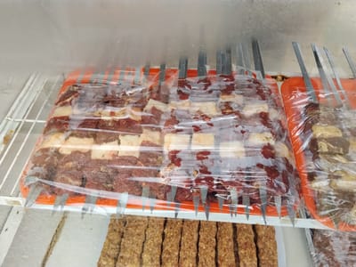 This image displays a close-up view of raw meat skewers, likely beef or lamb, packaged in clear plastic and arranged on orange trays. The skewers are made of metal and are partially visible poking through the plastic. They are stored on a white wire shelf inside a refrigerated unit, suggested by the condensation on the plastic packaging. Below the skewers, a tray of cooked, rectangular meat patties, possibly kebabs, is visible. The overall setting appears to be a food display or preparation area in a market or butcher shop in Ala-Buka, Kyrgyzstan. The lighting is bright, suggesting artificial illumination. There are no people visible in the frame.