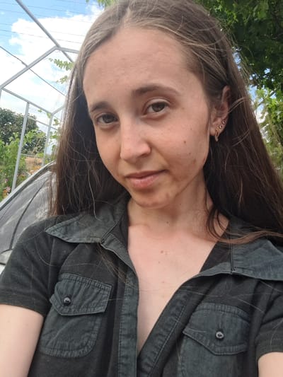 This is a selfie of a person in an outdoor setting, possibly a greenhouse or garden. The person has long, dark brown hair and is wearing a dark-colored button-up shirt. Their face is partially visible, with a hint of a smile. The background shows greenery and a structure that appears to be a greenhouse frame, with the sky visible through it. Sunlight suggests it is daytime. The image is tilted, giving a dynamic perspective.