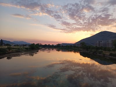 A wide-angle, landscape photograph captures a serene river scene at sunset in Trebinje, Bosnia and Herzegovina. The sky is painted with soft hues of orange, pink, and pale blue, with scattered, fluffy clouds reflecting in the calm water. In the foreground, the river stretches across the frame, its surface mirroring the dramatic sky and the surrounding landscape.
Along the left bank of the river, a collection of buildings and trees are visible, suggesting a residential or developed area. The middle ground features more foliage and structures, including a long, modern building with multiple windows, and further to the right, a series of multi-story apartment buildings with dark-trimmed windows. A steep, dark mountain rises prominently on the right side of the frame, its silhouette partially obscured by the setting sun. The overall impression is one of tranquility and natural beauty, enhanced by the warm, diffused light of the sunset.
