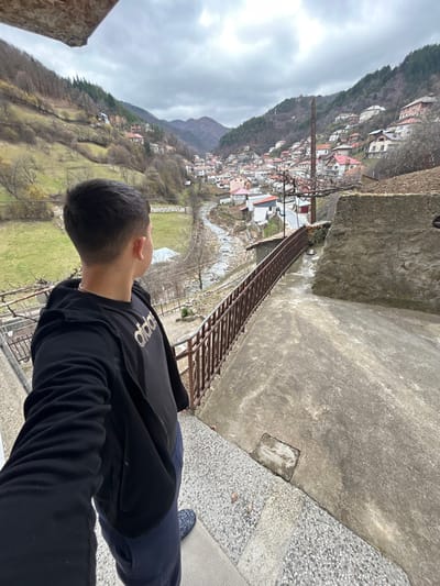 A male subject stands on a textured concrete platform, positioned with his back to the camera, looking down into a valley containing the village of Erma Reka, Bulgaria. The subject has short, dark hair and is dressed in a black zippered hoodie, a black t-shirt featuring a light-colored "Adidas" logo, dark blue sweatpants, and blue patterned slip-on shoes. To the immediate right, a brown metal railing descends along the edge of the platform, adjacent to a lower concrete surface and a rough-hewn stone wall. The valley below reveals a winding, low-water riverbed. Multiple multi-story houses with predominantly red and grey roofs are visible, built into the steep, partially vegetated hillsides. A narrow bridge with railings spans the riverbed in the midground. Sparse, leafless trees are scattered throughout the valley and on the lower slopes, while distant mountain ranges appear forested. The sky is uniformly overcast and grey, providing diffused natural light to the scene.