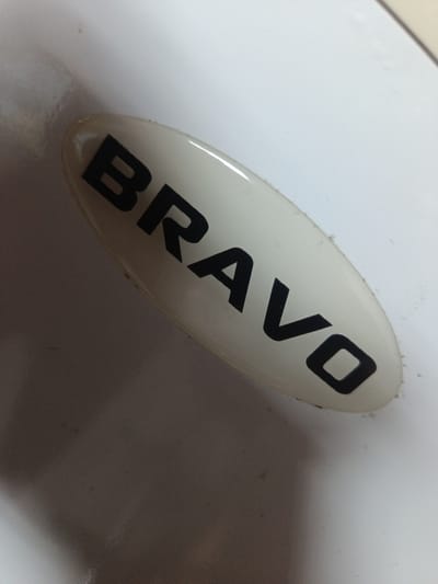 The image shows a close-up, angled shot of a white oval sticker with the word "BRAVO" printed in bold black letters. The sticker is affixed to a white, slightly textured surface, possibly a wall or an appliance. The lighting creates shadows that suggest the object is curved. The background is out of focus and appears to be a light gray or white. There are no people or identifiable landmarks from Zarubino, Russia, visible in the image.