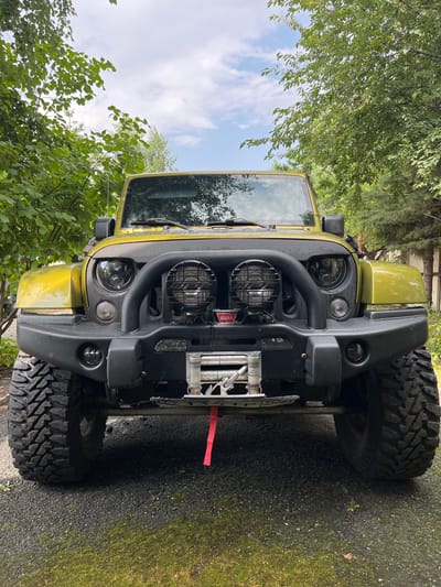 The image shows the front of a yellow Jeep Wrangler parked on a gravel surface outdoors. The Jeep has a black aftermarket bumper with a winch and two large circular lights mounted on it. It also has aftermarket headlights and large, aggressive off-road tires. The vehicle is surrounded by lush green trees and foliage. The sky above is partly cloudy. There are no people or animals visible in the image. The scene appears to be in a natural or semi-natural environment, possibly a driveway or a rural setting. The specific location in Старомалиновка, Russia is not determinable from the visual information provided in the image.