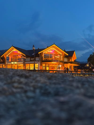 The photograph captures a building at dusk in Zakopane, Poland, with the sky a deep blue, transitioning from twilight. The building, illuminated by warm orange and pink lights, features a distinctive architectural style with prominent wooden elements and steep, gabled roofs adorned with decorative trim. Large windows reveal the interior, where tables and chairs are visible. A sign reading "GRILL-PIZZI" is prominent on the facade. A blue parking sign is also affixed to the exterior. Several individuals are seen standing and walking outside the building, along a wooden railing that separates them from the viewer. In the foreground, a rough, textured surface, possibly stones or rocks, is out of focus, creating a depth-of-field effect. The scene suggests a lively evening atmosphere in a tourist area of Zakopane, known for its mountain resort ambiance.