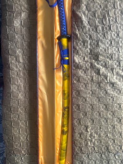 The image displays a decorative katana sword presented in an open box. The box is lined with orange satin and rests on a textured brown surface, possibly a blanket or upholstery. The katana's handle is wrapped in blue cord with silver accents, and the scabbard is yellow with a marbled pattern and an illustration of a woman holding a fan. The scene is lit by natural sunlight, casting shadows and highlighting the textures. There are no people visible in the image. The location in Stepnogorsk city administration, Kazakhstan is not determinable from the provided image.