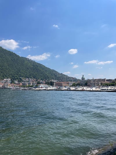 The image shows the city of Como, Italy, from a high vantage point near the water. The foreground is the water of Lake Como, showing slight ripples. Moving towards the left, the city climbs a green, tree-covered hillside. Buildings of varying sizes and colors are interspersed among the trees. At the top of the hill, the city continues along the water’s edge with taller buildings and a prominent dome. The sky above is blue with scattered, puffy white clouds.