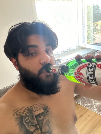 A man with dark hair and a beard is drinking from a green glass bottle with a Beck's beer label. He has a tattoo on his chest of a lion and praying hands with a cross and a crown of thorns. He is indoors, in front of a window with sheer white curtains. The environment appears to be a home with wooden flooring and a patterned rug visible. There is no indication of the city of Mainburg, Germany in the image.