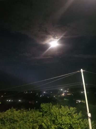 This image depicts a nighttime scene in Radishevo, Bulgaria, dominated by a bright moon illuminating a cloudy sky. The moon's intense light creates a halo effect and flares extending outwards. Below the sky, a landscape unfolds, featuring a silhouette of trees in the foreground, obscuring the lower part of the image. Beyond the trees, scattered lights indicate buildings or streetlights in a town or village area. A utility pole with wires stretches vertically along the right side of the frame, also illuminated by the moonlight. The overall atmosphere is dark and atmospheric, characteristic of a moonlit night.