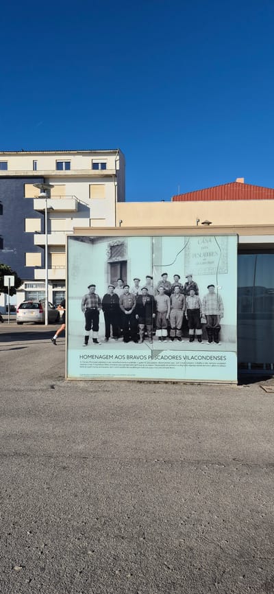 A wide, low-angle shot captures a mural displayed on a building in Vila do Conde, Portugal. The mural, a black and white photograph, features a group of seventeen men, presumably fishermen, standing in two rows. The men are dressed in dark trousers, shirts, and some wear berets. The mural is mounted on a light-colored facade. Above the mural, a modern, multi-story building with dark and light blue sections is visible. To the right of the mural, a section of a building with a red-tiled roof is present. The foreground consists of a paved area, likely a street or sidewalk. A person is seen running on the left side of the image, their body blurred due to motion. The sky is a clear, bright blue.