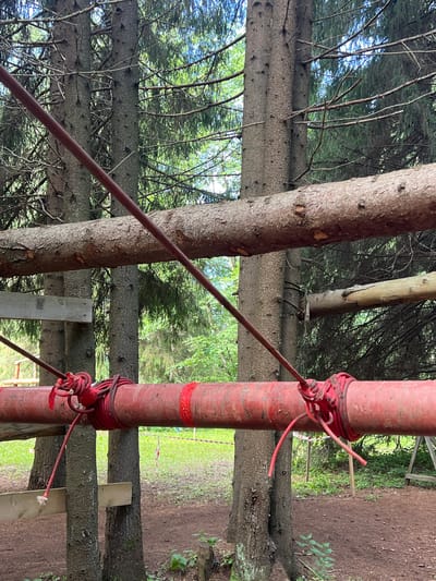 In the provided image, a section of an adventure park obstacle course is visible, set within a forested area. Two mature pine trees serve as natural supports for a horizontal red pipe, which is secured by thick red ropes. Above this, a natural log is also suspended horizontally between the trees, likely part of another element of the course. The ground below is covered in loose dirt and some sparse vegetation, with a distant glimpse of a grassy area marked by red and white caution tape, indicating the boundaries of the park. The overall scene suggests a rugged, outdoor recreational environment in the city of Sergino, Russia, with no people or animals present in the frame.
