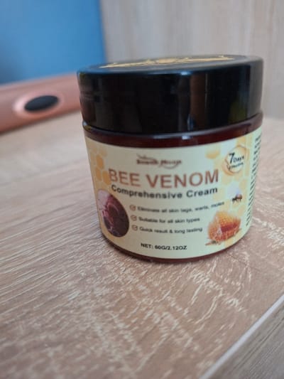 A dark amber-colored jar with a black screw-on lid, labeled "BEE VENOM Comprehensive Cream," is positioned prominently on a light-colored wooden surface. The label features a honeycomb pattern background and includes text stating "Seventh Heaven," "7days effective," and "NET: 60G / 2.12OZ." Bullet points indicate the cream can "Eliminate all skin tags, warts, moles," is "Suitable for all skin types," and provides "Quick result & long lasting." Illustrations on the label depict a dark red irregular mass, a bee, and a honeycomb section. The wooden surface displays distinct linear grain patterns. In the blurred background, to the left of the jar, a rose-gold colored object with a black oval feature is partially visible. Further back, a blue surface is also out of focus. The image contains no visual information to determine its specific location within the city of Selyanin, Bulgaria, or any other geographical area.