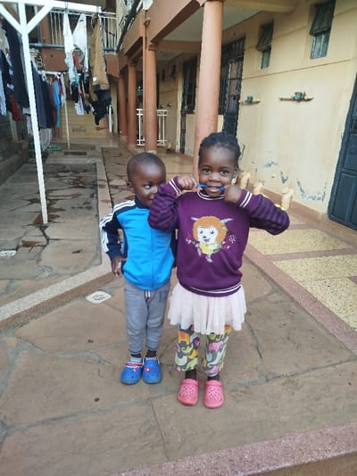 Two young children, a girl and a boy, are captured standing on a paved outdoor surface, likely in a residential courtyard in Kihara ward, Kenya. The girl, on the right, wears a dark purple long-sleeved top featuring a cartoon character, a light-colored tutu-style skirt, multi-colored patterned leggings, and pink Crocs-style shoes. She holds a blue toothbrush to her mouth, smiling brightly at the camera. The boy, standing to her left, is dressed in a blue and white long-sleeved jacket, grey pants, and blue Crocs-style shoes; he is looking away to the left with a subtle smile.

In the background, there are multi-story residential buildings with light brown or beige facades and dark-framed windows and doors. Brown support pillars are visible, indicating covered walkways or balconies. To the far left, a white clothesline structure is laden with various clothes hanging to dry. The ground is paved with flagstone-like tiles in shades of brown and grey, and small circular grates are embedded in the pavement. The natural light suggests it is daytime, possibly an overcast or early/late hour given the soft lighting. The children appear happy and playful, posing for the photograph. No legible text is visible in the image.