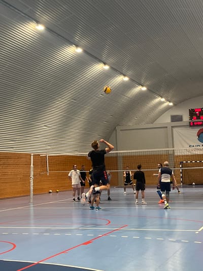 The scene depicts a handball game or practice indoors, likely in Pobrdje, Montenegro based on the presence of a scoreboard with 'FAIR PLAY Sports' visible. Several people are present, with one individual in mid-air, appearing to spike or hit a yellow handball. Other players are positioned around the court. The location is a gymnasium with a blue playing surface, a net dividing the court, wood paneling, and a corrugated metal roof with overhead lighting. A scoreboard is visible in the background.